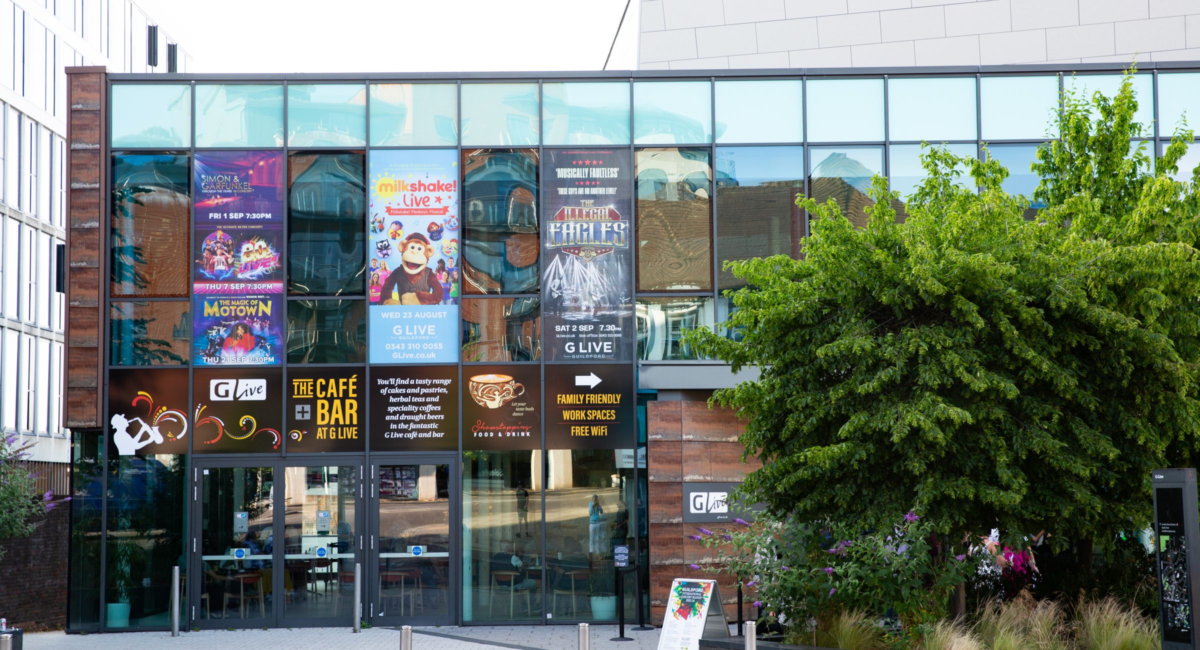 An exterior shot of the building with coloured posters wrapping some windows, a sign outside and green pruned large trees.