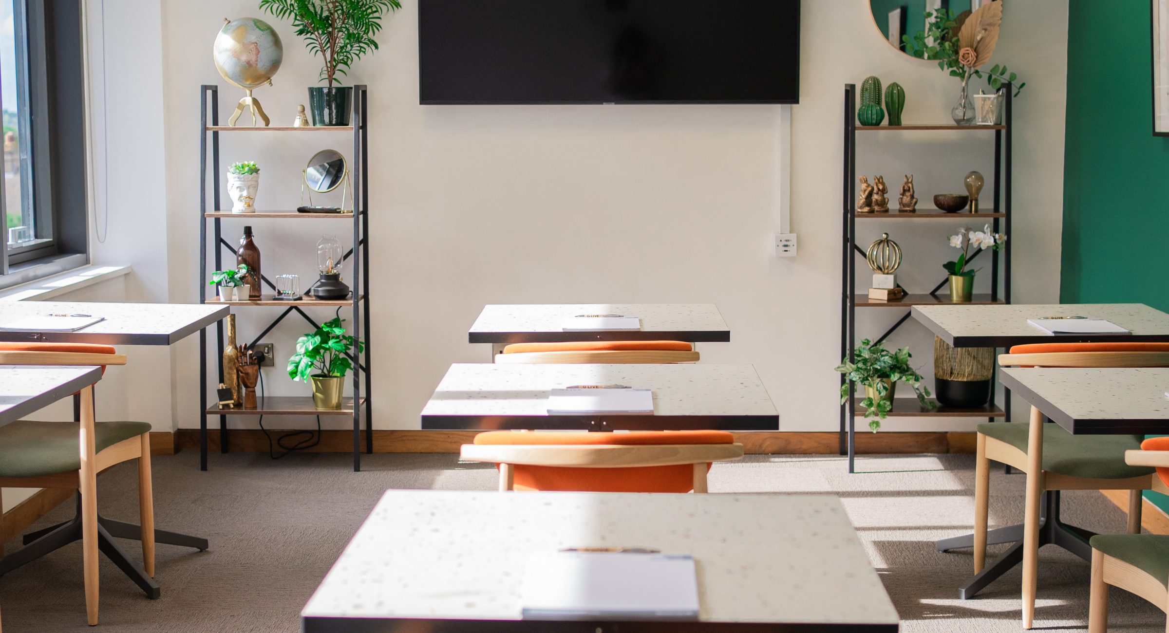 A centred shot of a row of marble tables and orange/green chairs focused on a tv screen and shelves dressed with greenery and knick knacks.