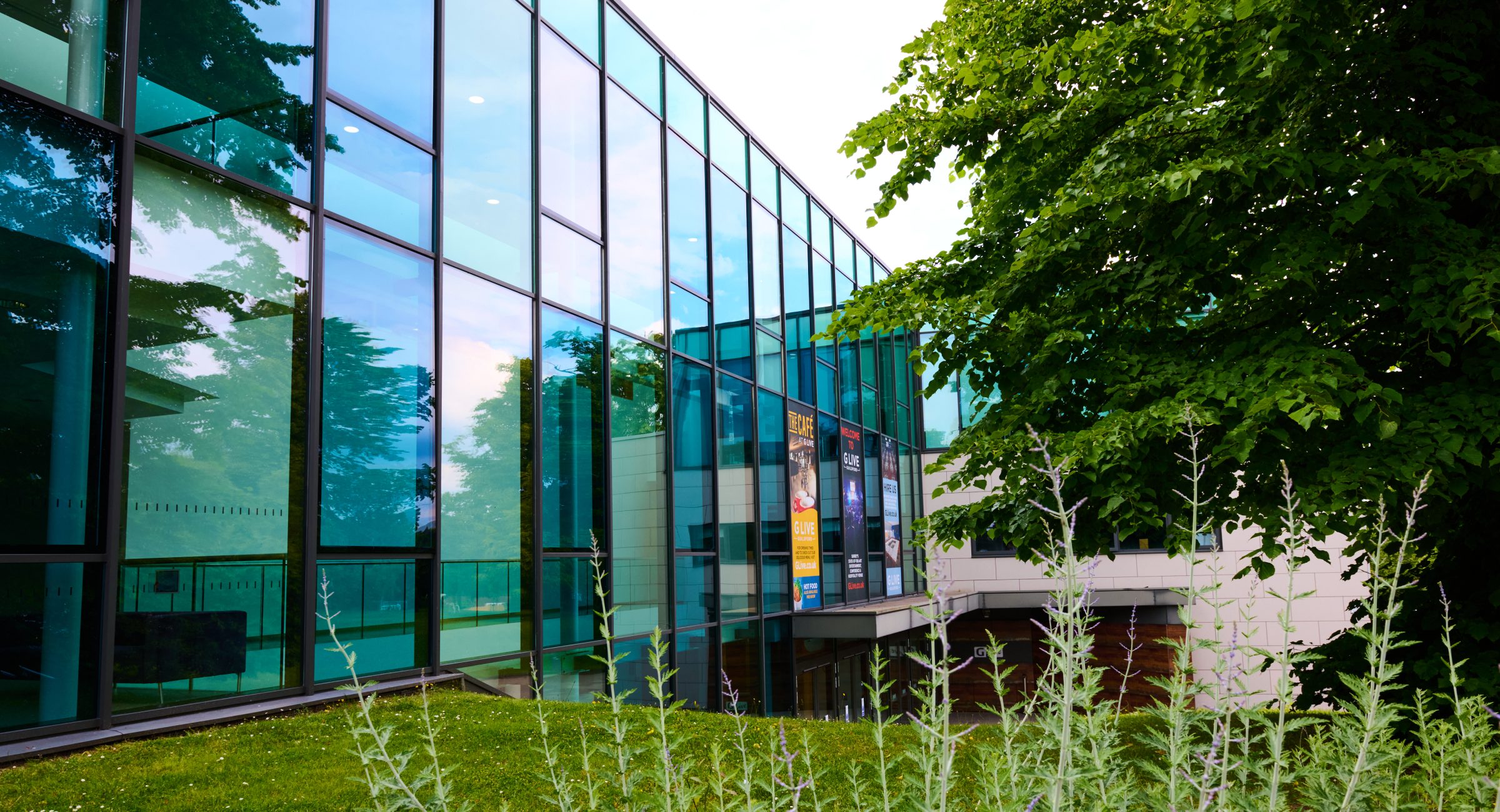 An exterior shot of the gleeming glass walls of G Live with green foliage and trees in the foreground.