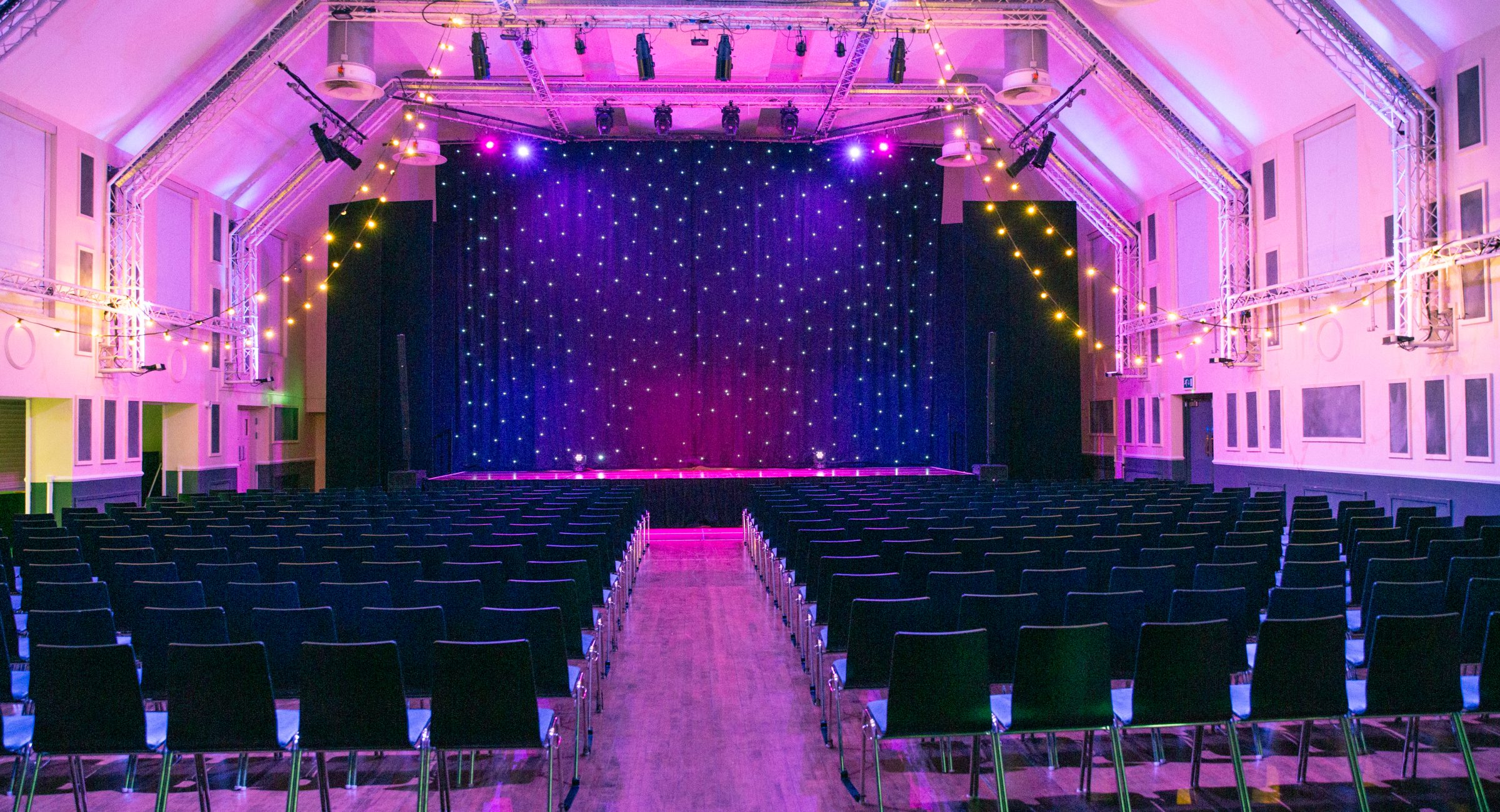A pink washed room with chairs in rows facing a star curtained stage and festoon lighting hanging from the stage truss lighting rig.