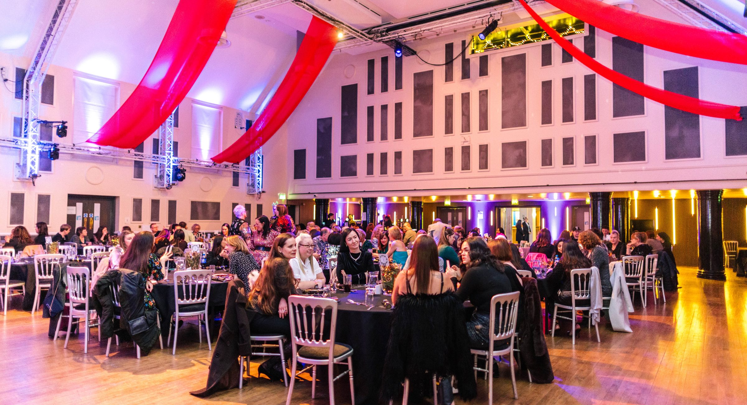 Seated guests in a large room on silver chairs around round tables, with red material hanging from the canopy.