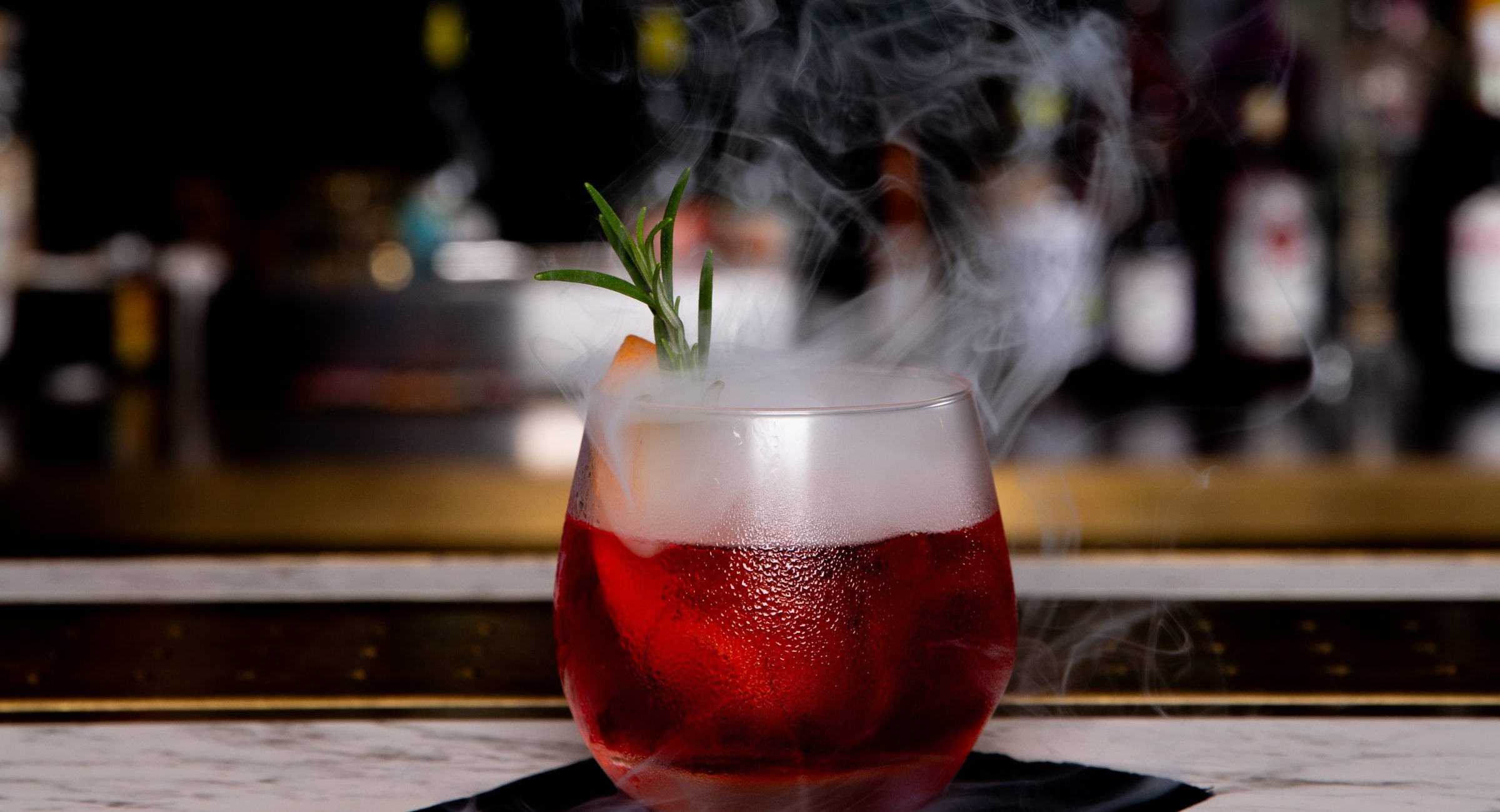 A smoking negroni on a black napkin on a marble bar with blurred bottles in the background.
