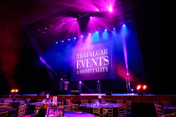 A room lit in pink and blue with a screen onstage displaying Trafalgar Events & Hospitality and tables with silver chairs around them.