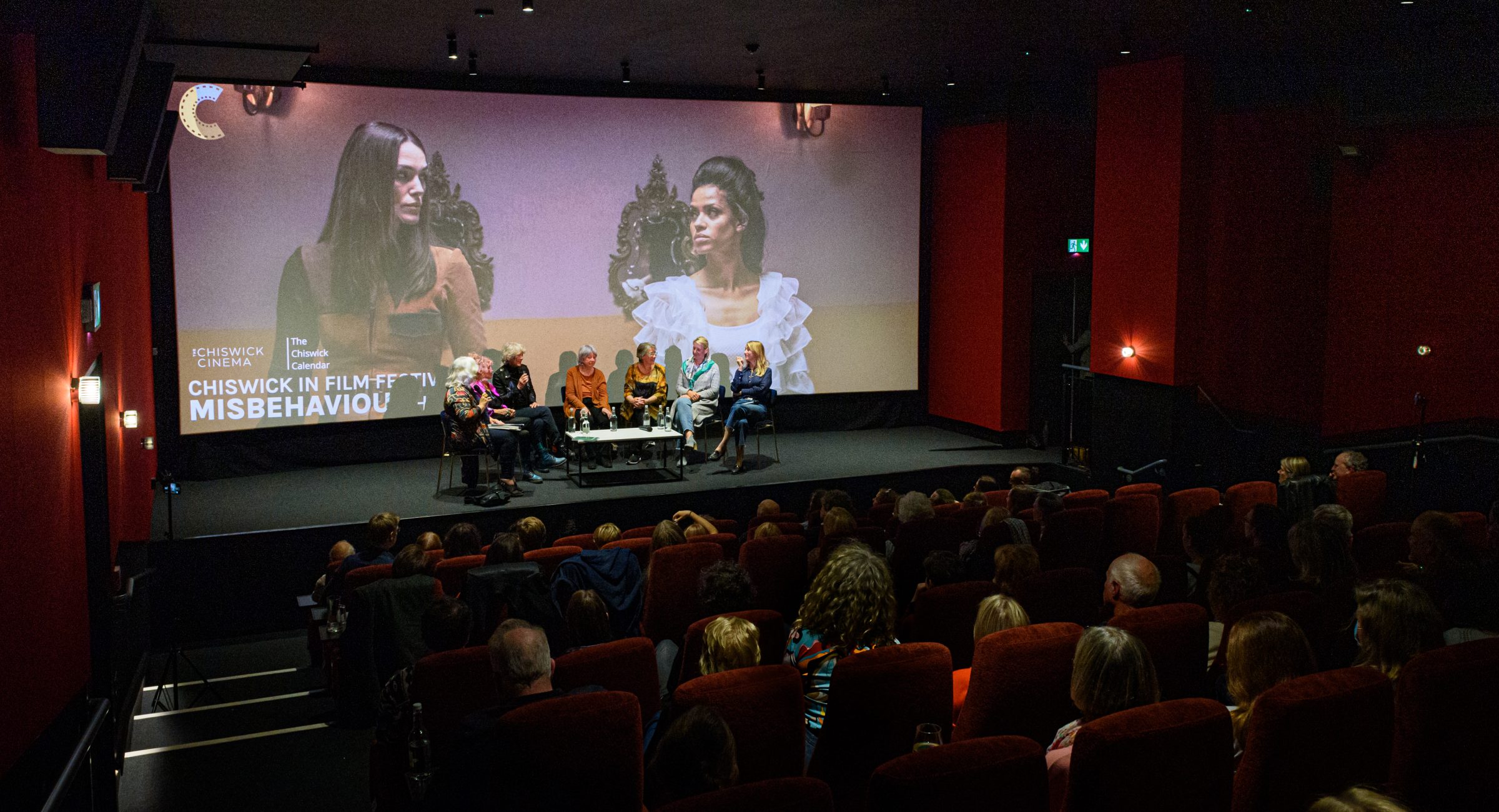 Audience member sat in dark red cinema seats looking at a seated panel of people on stage in front of a large cinema screen.