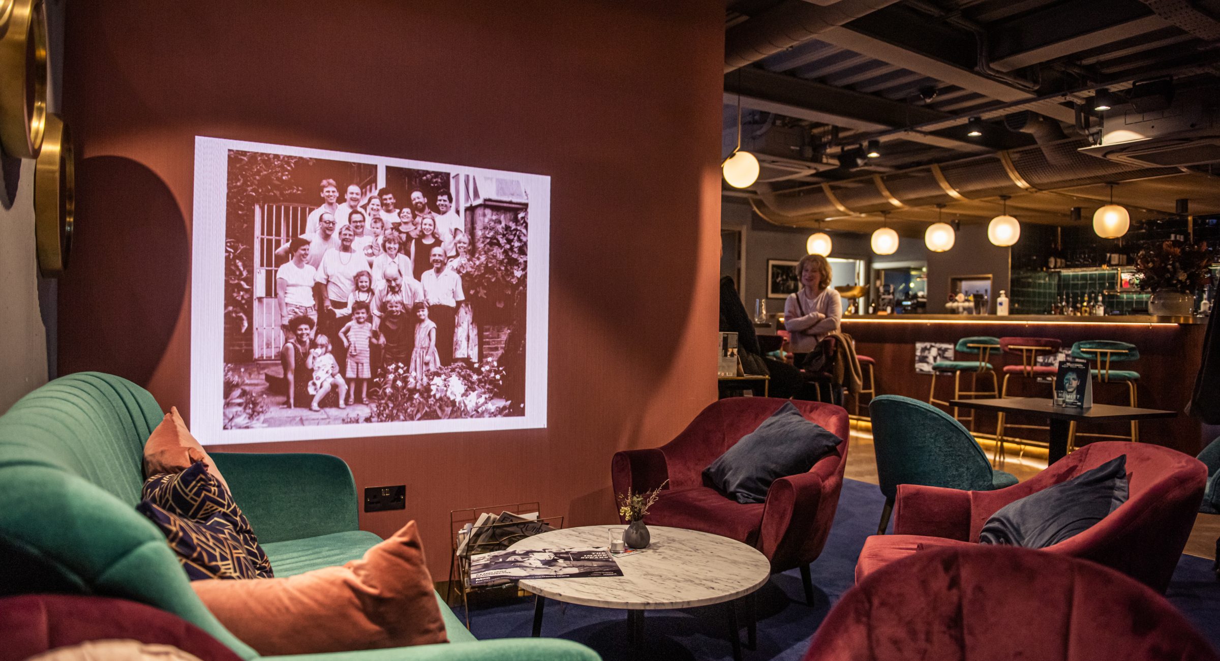 Colourful purple and blue armchairs with throw pillows on and a projected black and white image on a terracota wall with a bar in the background.