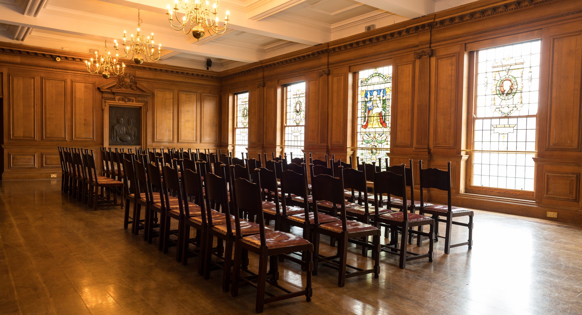 A wood panelled room with chandeliers and silver backed chairs in rows facing a microphone.