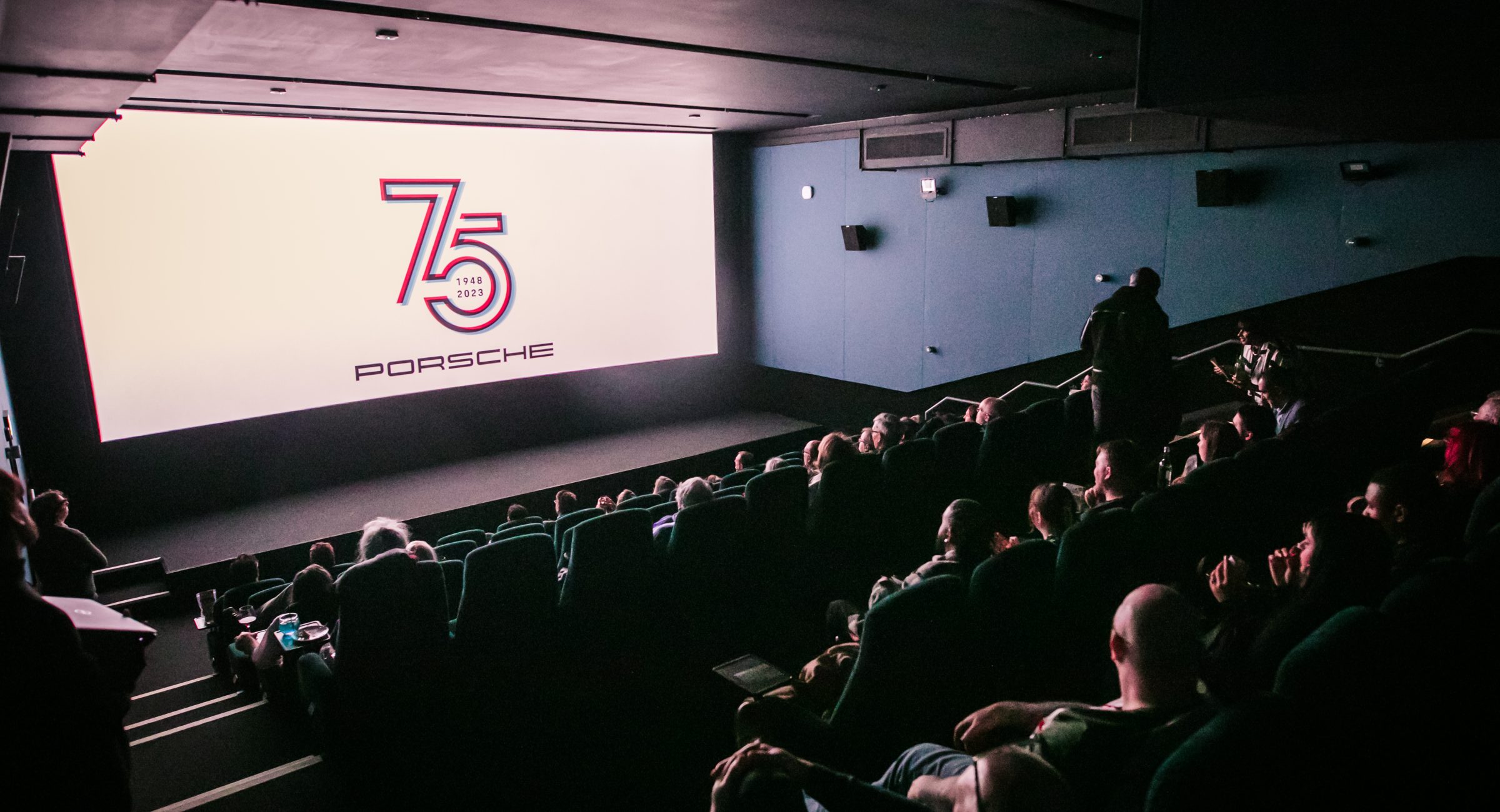 A full screen of guests attending a 75 porsche event with this projected on the screen.